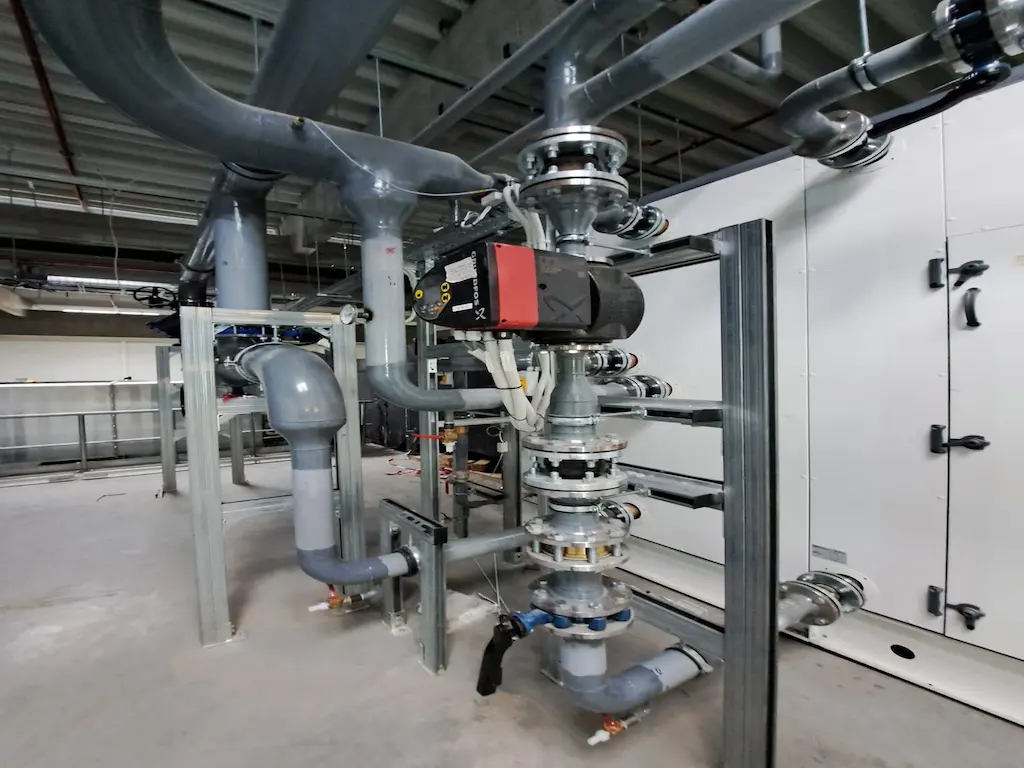 A Heat Builder Kft. által telepített ipari gépészeti központ részlete, központban egy professzionális Grundfos keringető szivattyúval, szürke csővezeték-hálózattal és nagy teljesítményű légkezelő egységgel a háttérben.