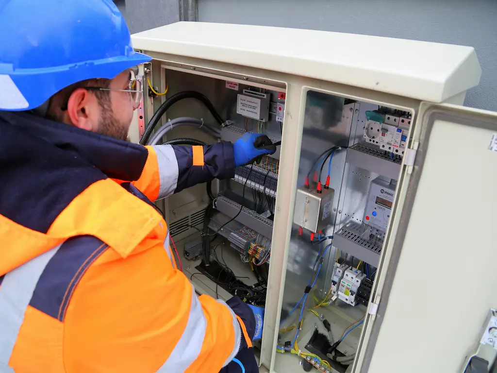 A Heat Builder Kft. villanyszerelő szakembere kék védősisakban és láthatósági ruházatban végzi egy kültéri elektromos vezérlőszekrény precíziós kábelezését és beüzemelését.