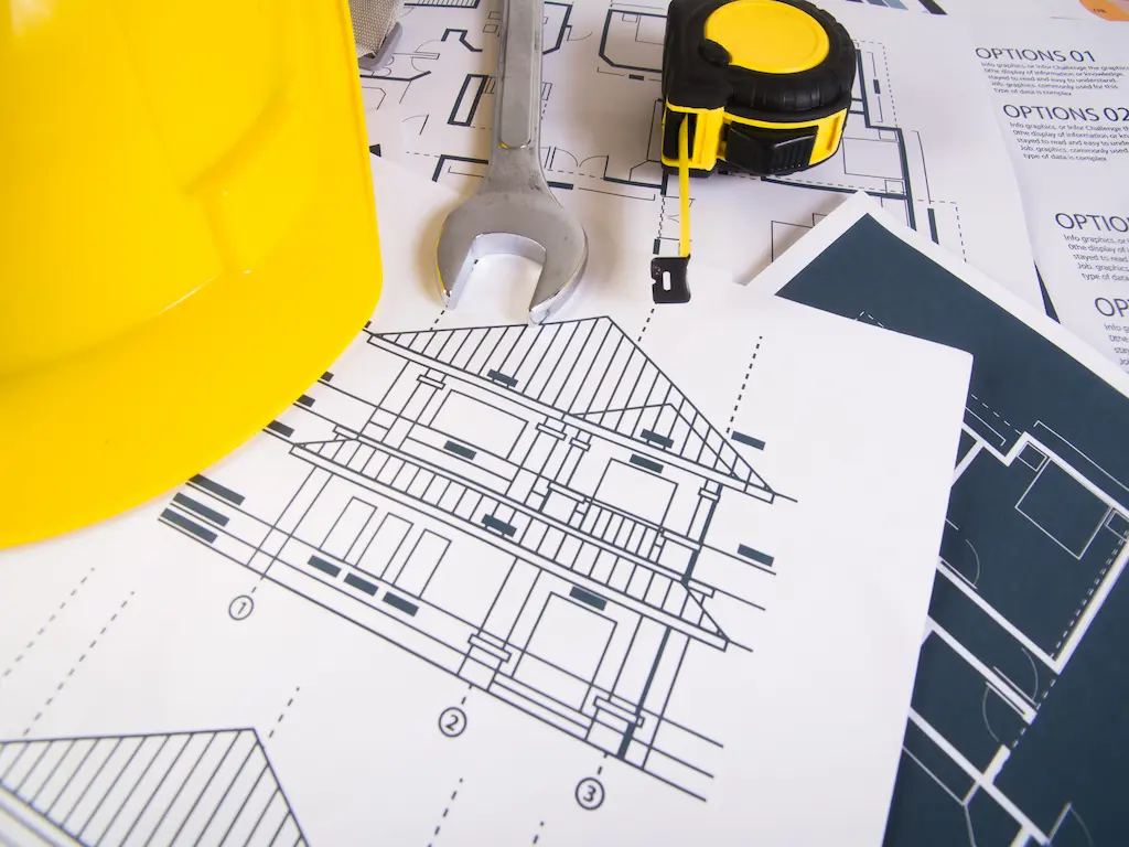 Részletes épületgépészeti tervrajzok, mérőszalag, villáskulcs és egy sárga védősisak az asztalon, amelyek a Heat Builder Kft. precíz mérnöki előkészítését szimbolizálják.
